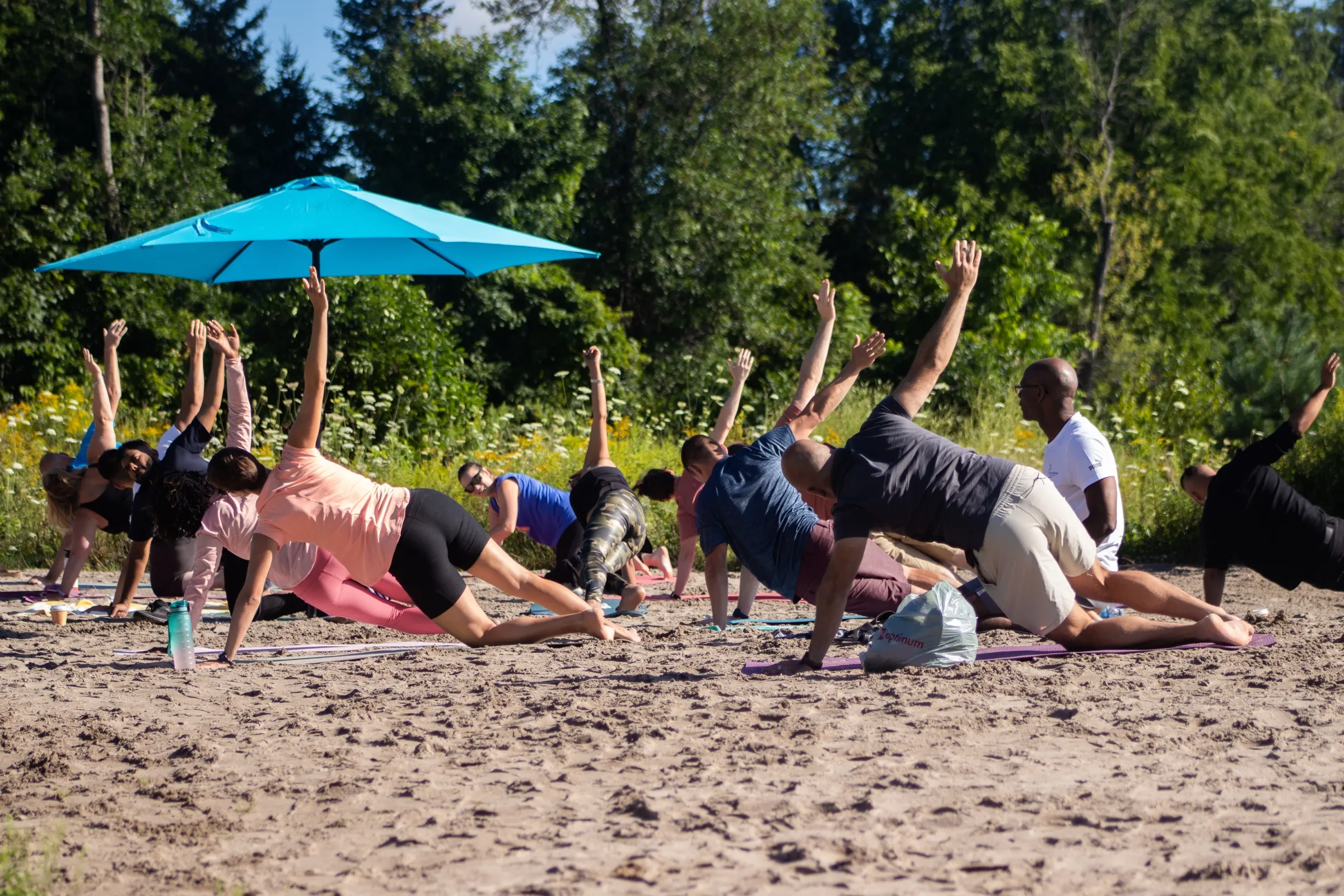 A yoga class on the sand, a dozen people in a side stretch on mats under a blue umbrella, with trees and wildflowers behind.