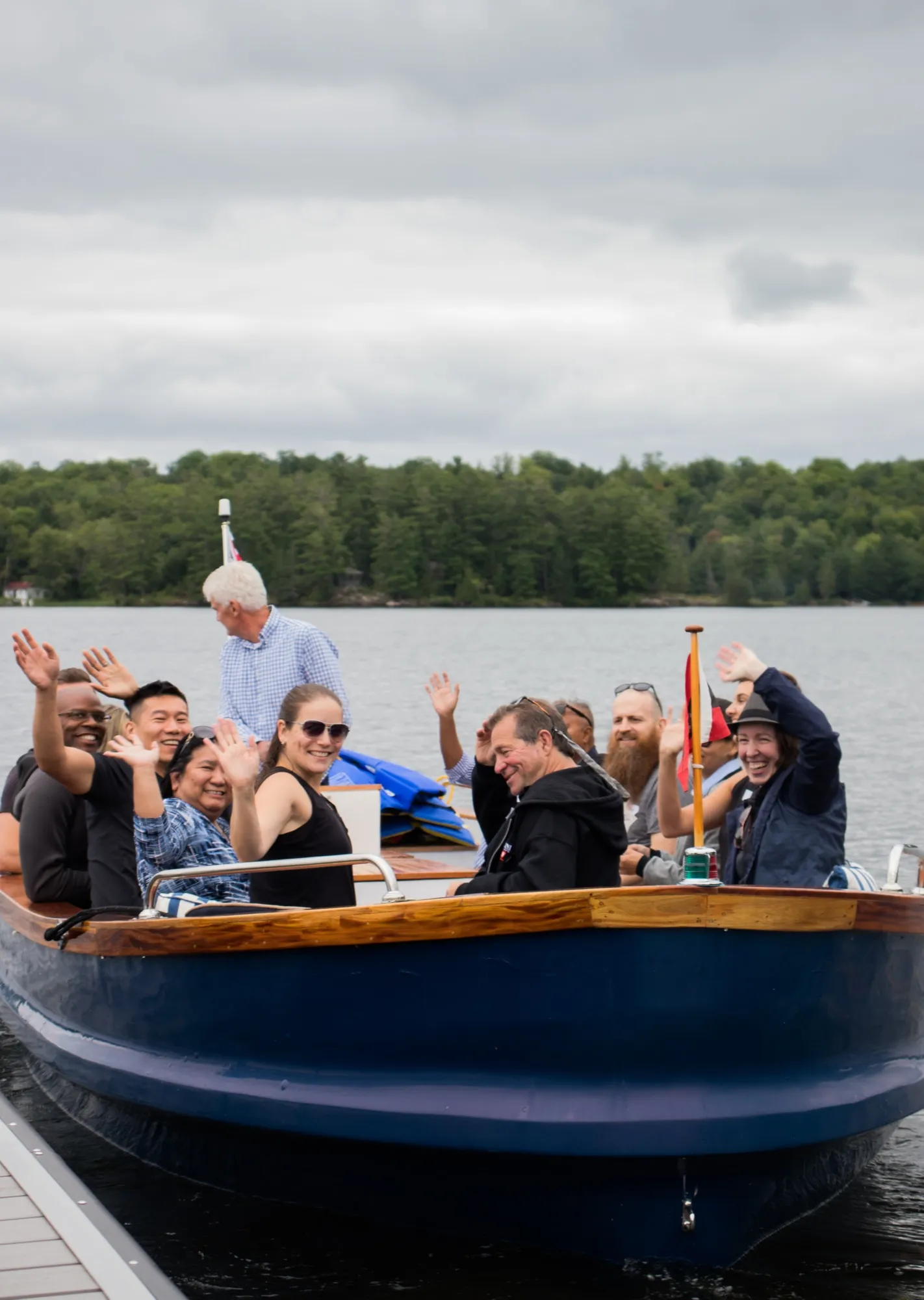 Guests aboard a navy and varnished wooden boat at the dock, waving and laughing, with the far shore behind them.