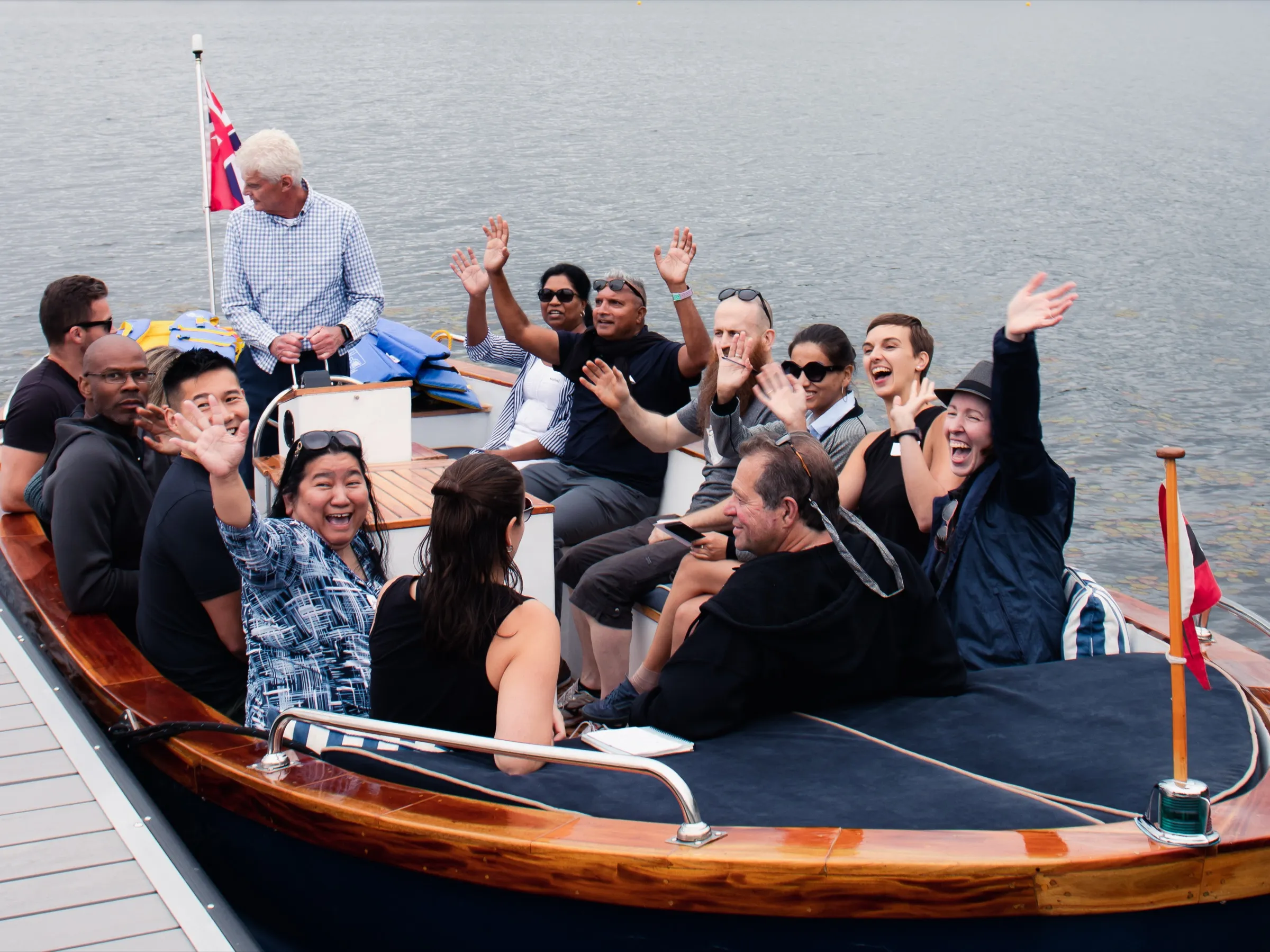 A dozen people aboard a varnished wooden boat on the lake, laughing and waving at the camera.