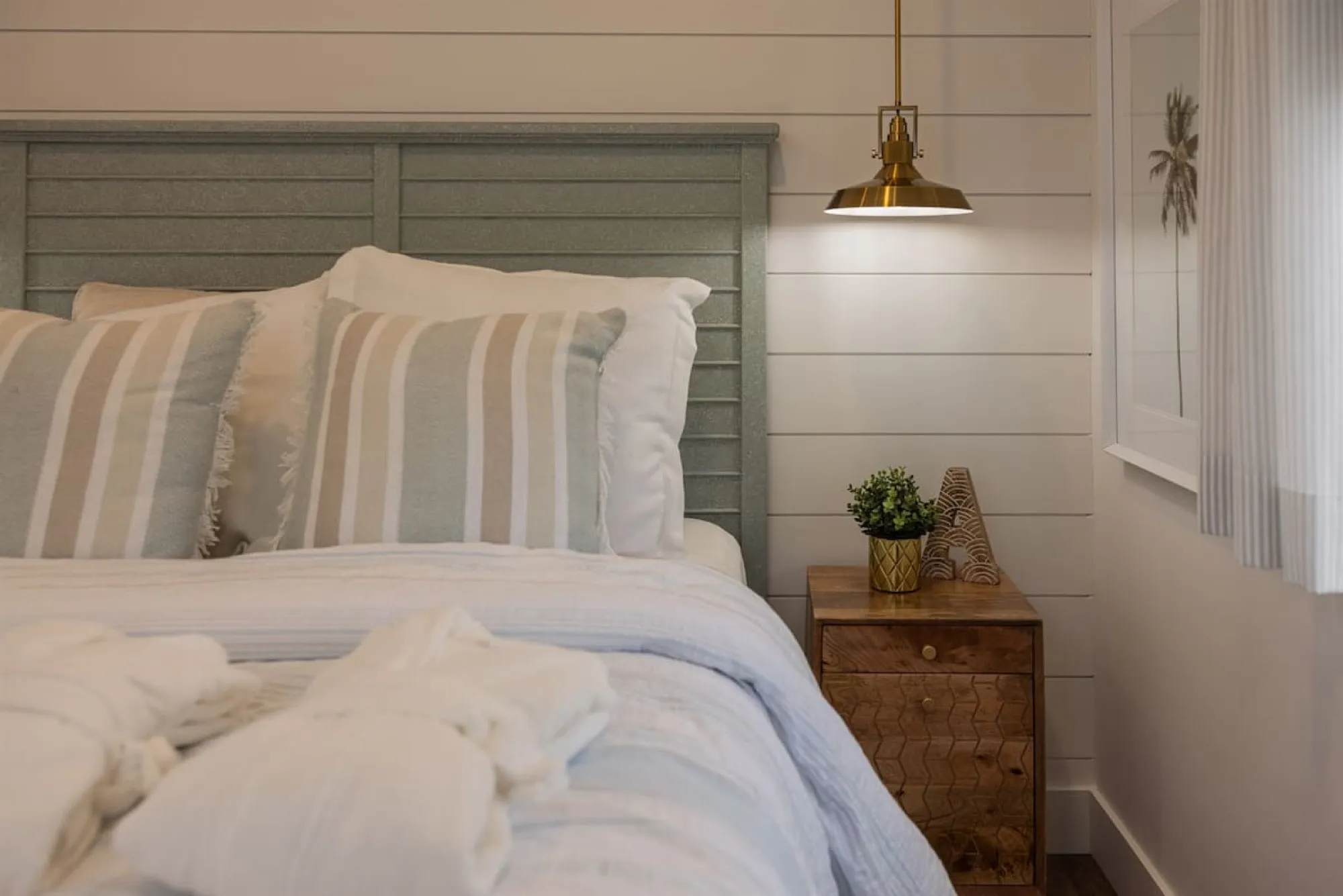 The Beach bedroom, with a sage-green slatted headboard, striped and fringed cushions, and a brass pendant light over a wooden nightstand.