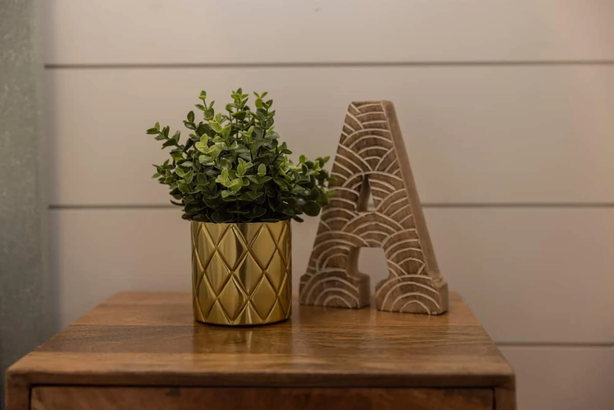 A potted plant in a gold faceted pot beside a carved wooden letter A on the nightstand in The Beach.