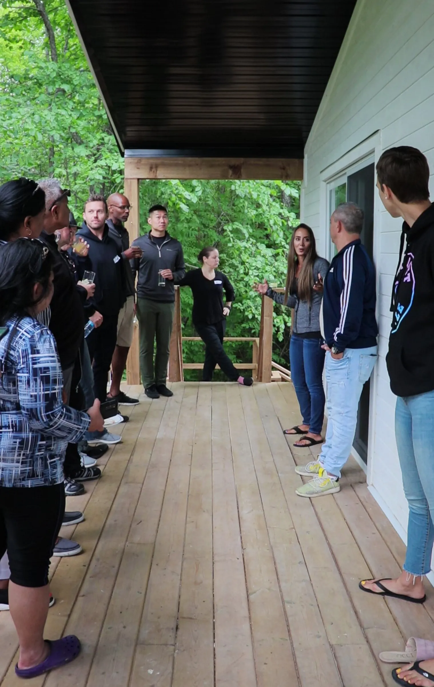 A group standing along a cabin deck with drinks and name badges, listening to a host speaking to them.