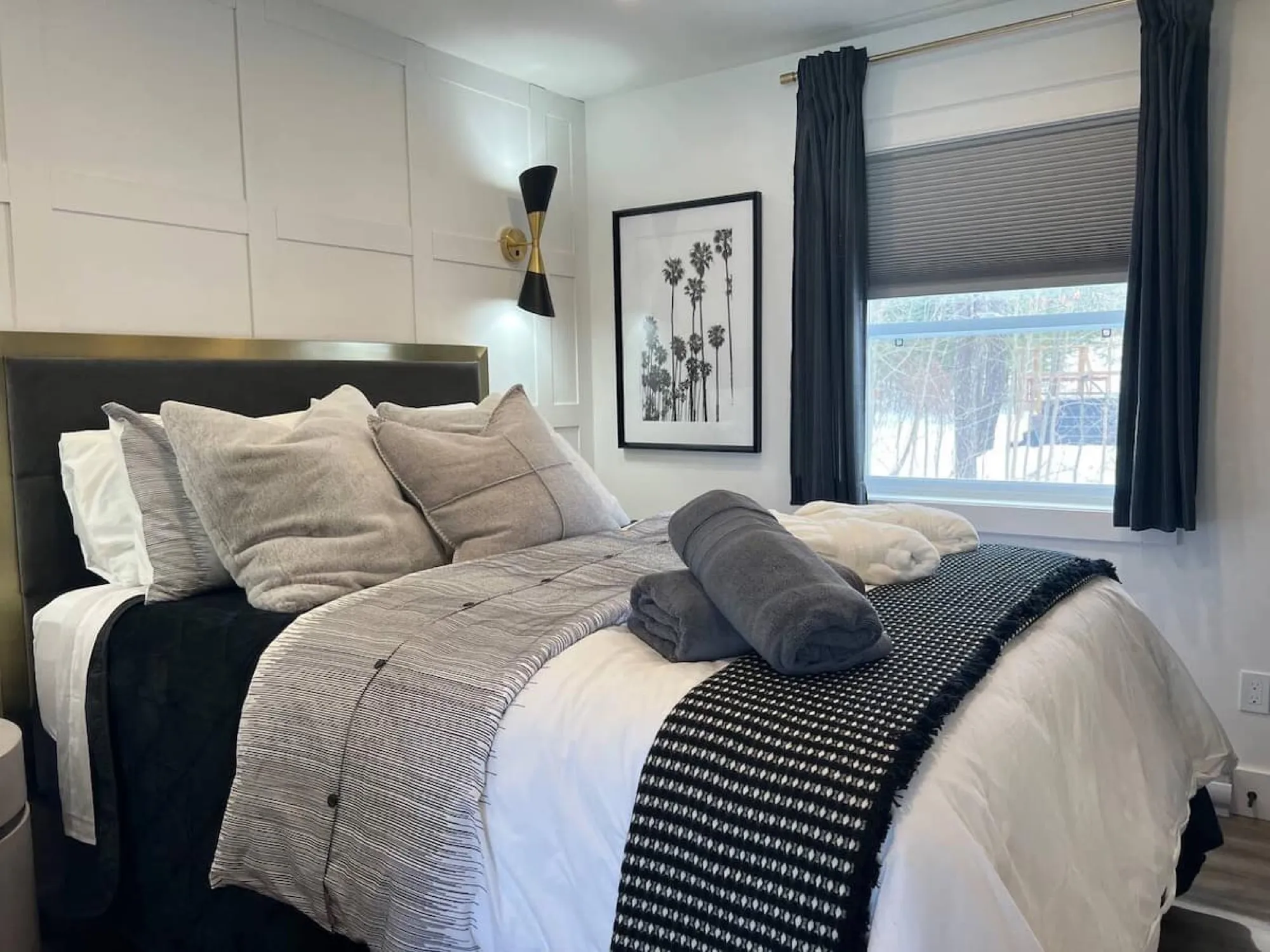 The Vintage Hollywood bedroom, with a brass-trimmed charcoal headboard, layered grey bedding and a black-and-white palm-tree print beside a curtained window.