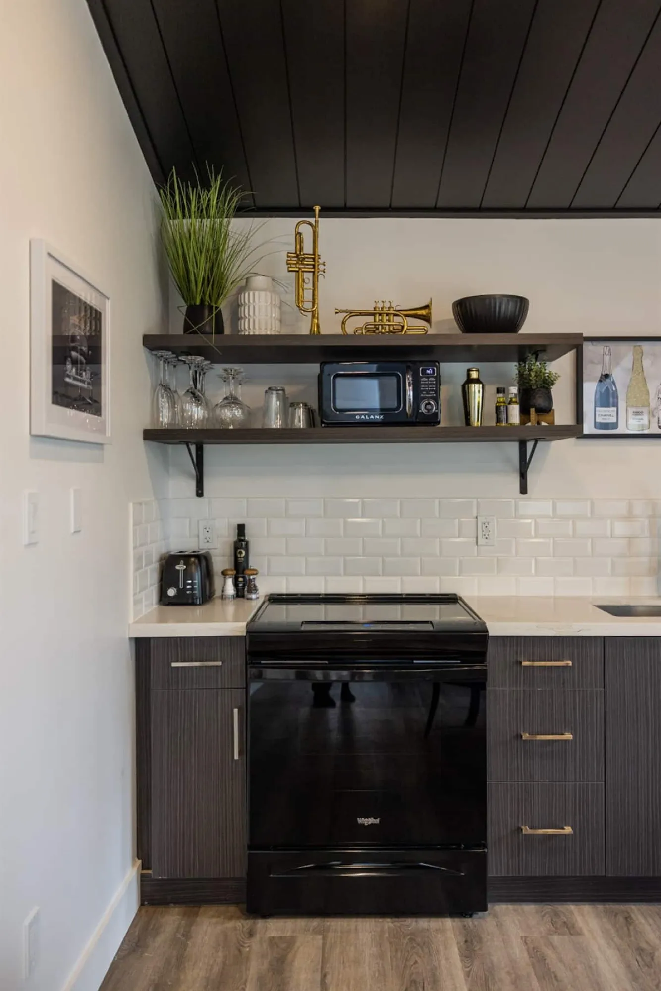 The Vintage Hollywood kitchen, with dark cabinetry, a white subway-tile backsplash and open shelves holding glassware and brass horns under a black plank ceiling.