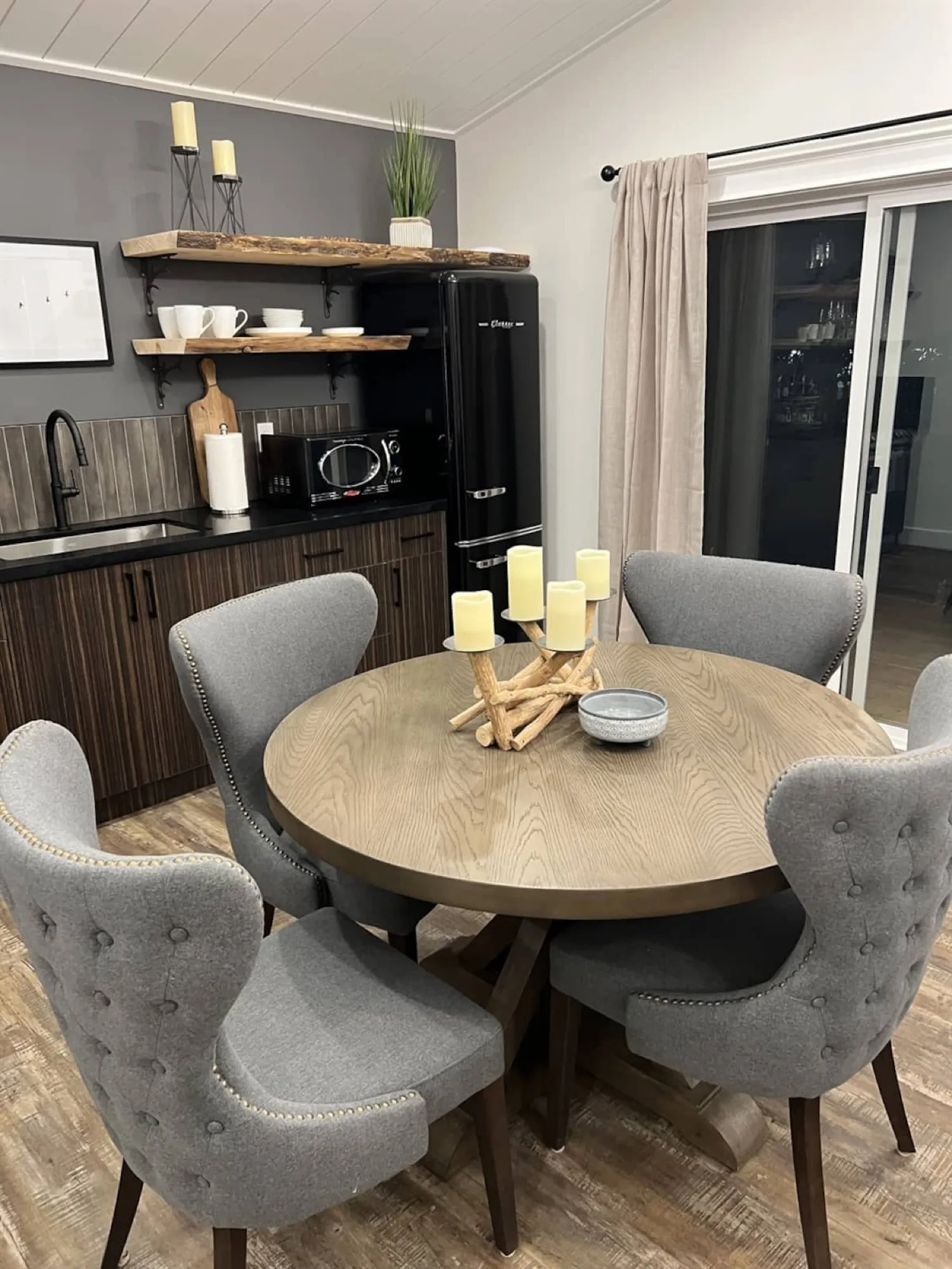 The Rustic Lodge dining area, with a round oak table and grey upholstered chairs, live-edge shelves and a black retro fridge.