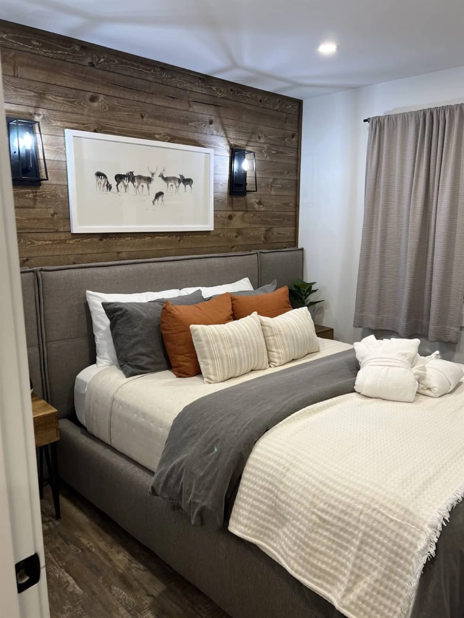 The Rustic Lodge bedroom, with an upholstered bed against a barnboard wall, a framed deer photograph and black lantern sconces.