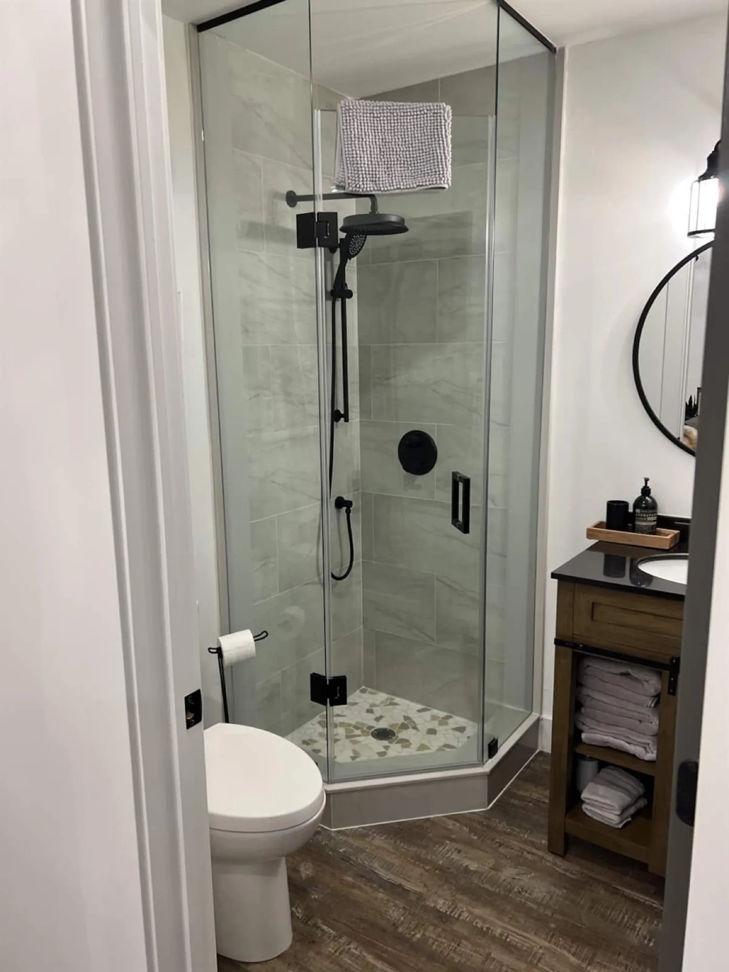 The Rustic Lodge bathroom, with a glass corner shower over pebble-mosaic tile, black fixtures and a wood vanity holding folded towels.