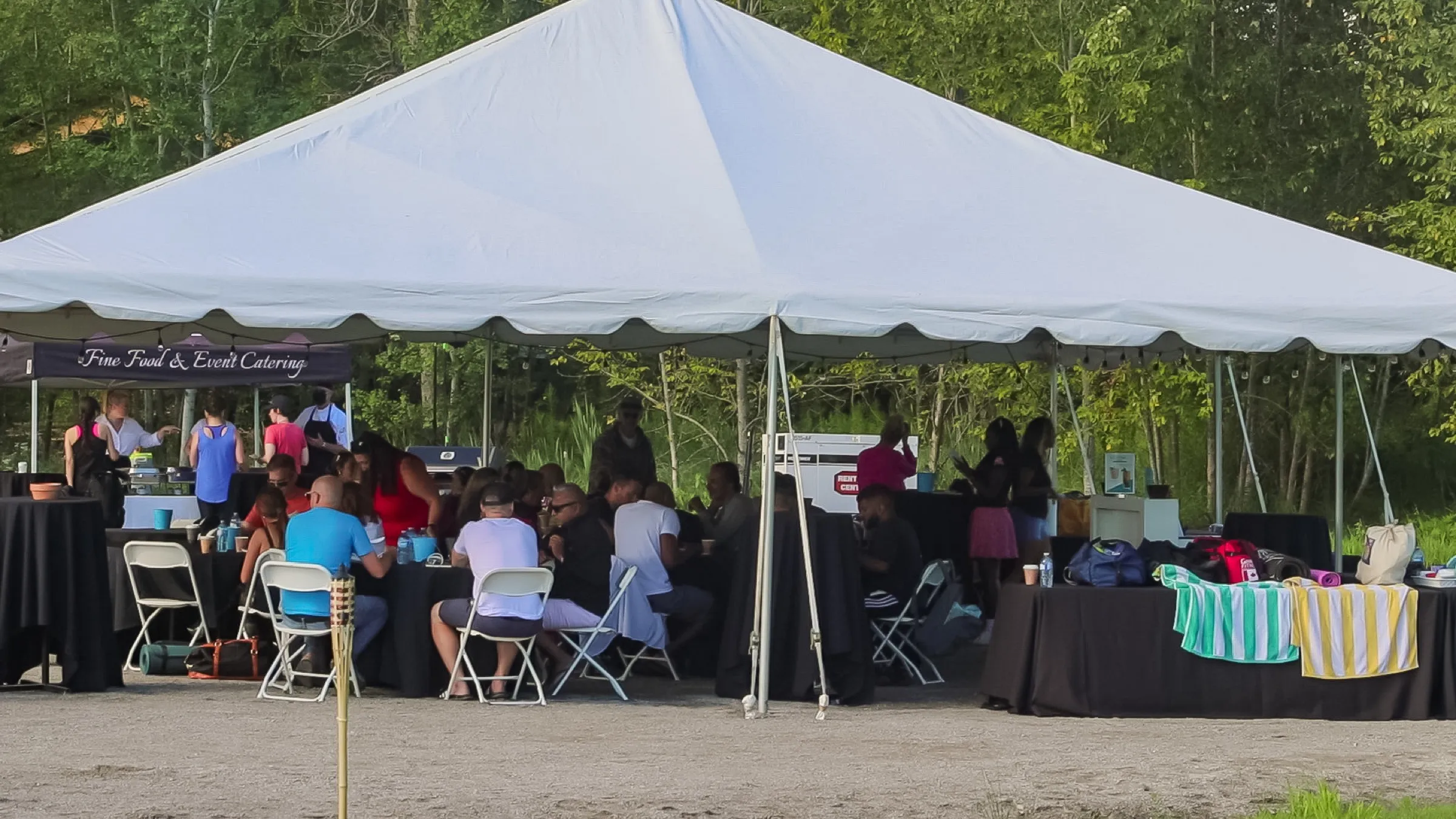 A large white marquee on a summer afternoon with guests seated at draped tables, a catering stand at one end and towels and yoga mats stacked on a table.