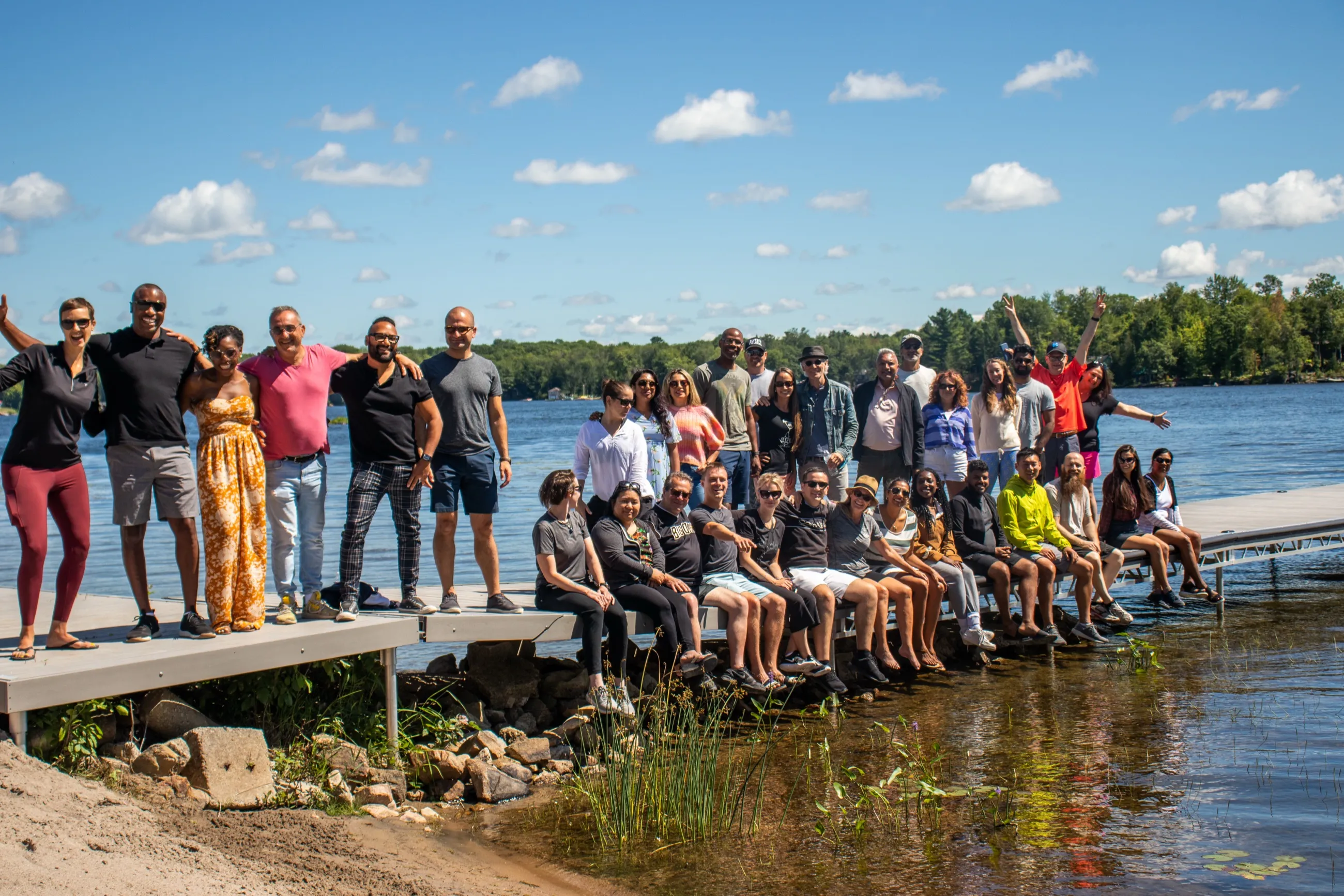 A large group of guests together on the dock at Inspire Beach Resort, some standing with arms raised and others sitting with their feet in the lake, under a bright sky.