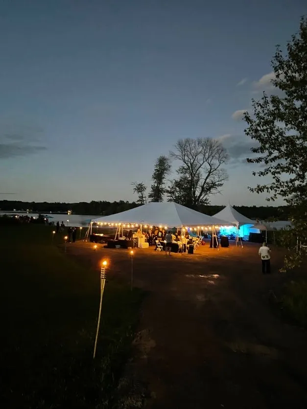 Two white marquees lit from within at dusk beside the lake, with bamboo torches lining the path to them.