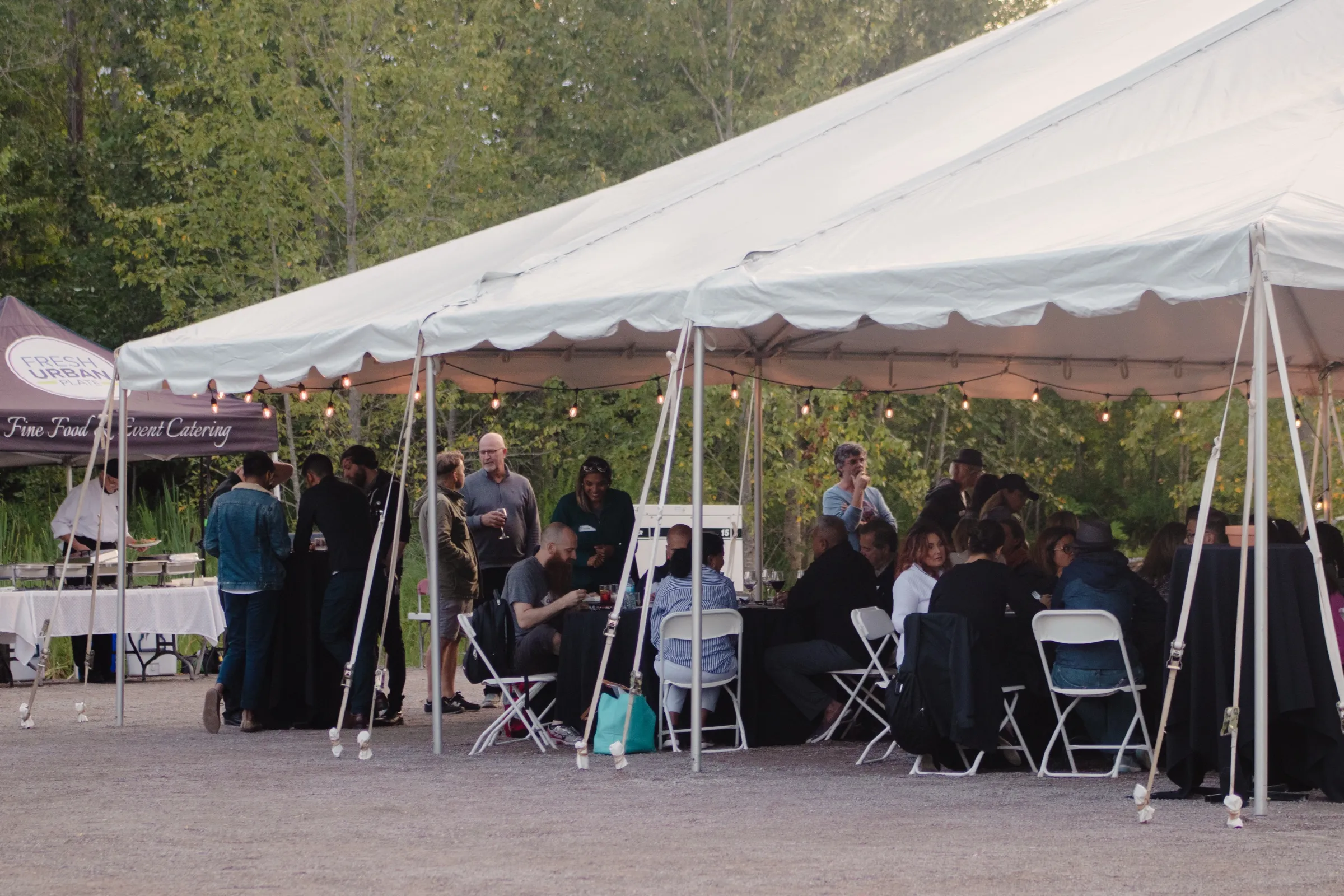 Guests dining at long tables under a white marquee strung with festoon lights at dusk, with a catering stand at one end and forest behind.