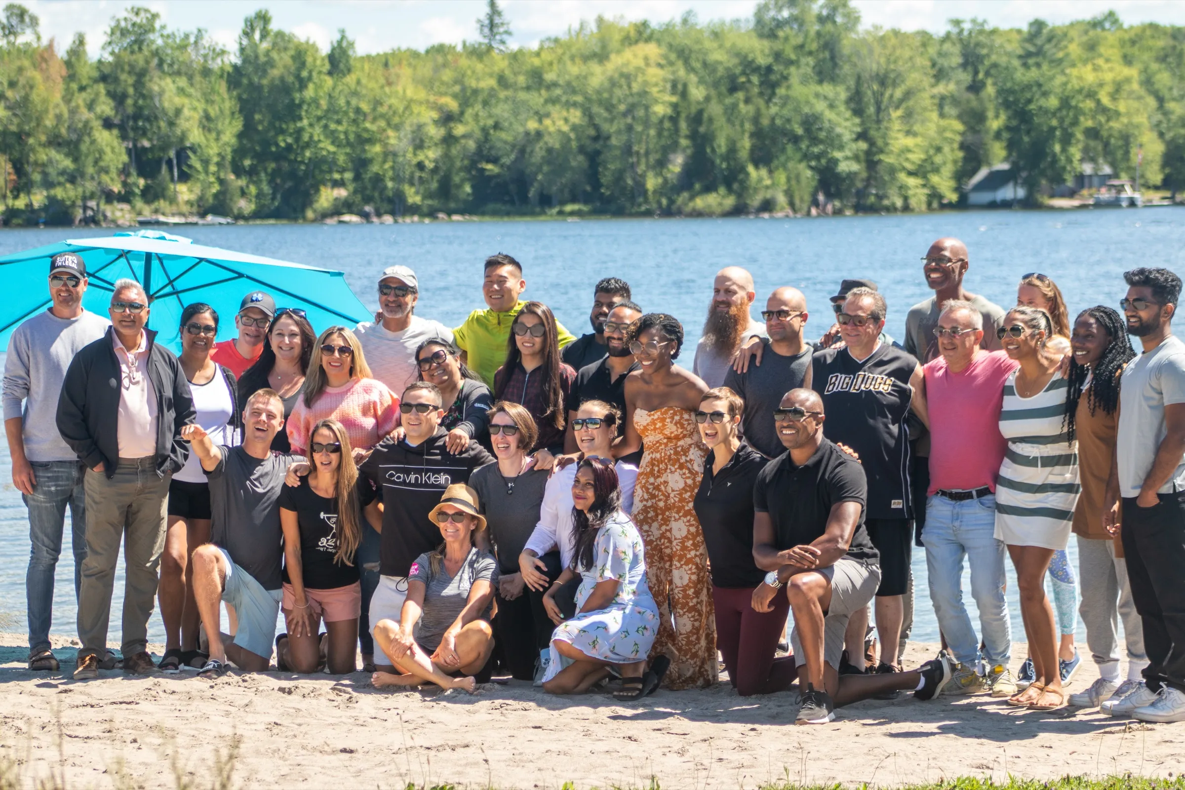 A group of about thirty people posed together on the sand in front of the lake on a bright day, with the far shore behind them.
