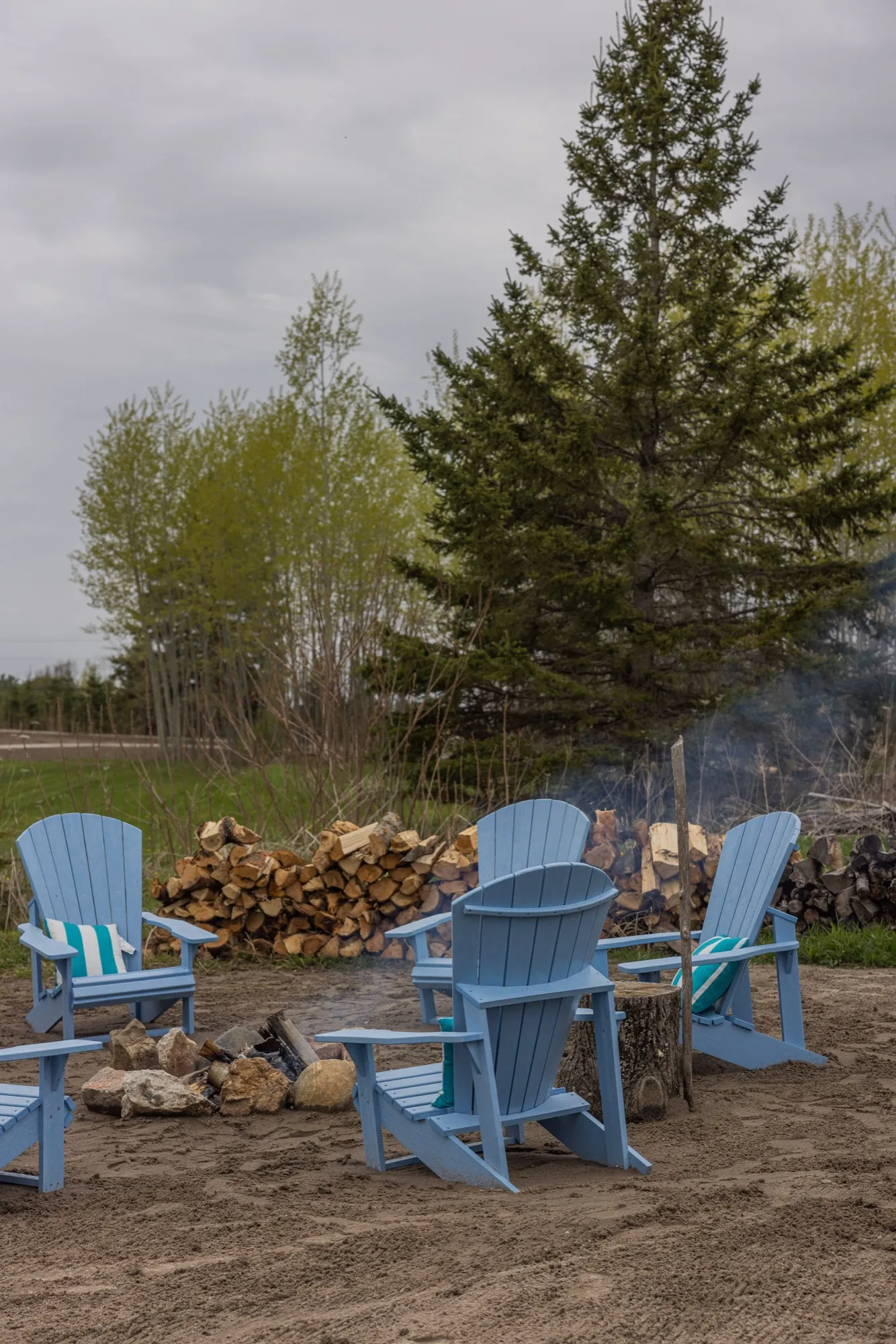 Blue Adirondack chairs set around a stone fire ring on the sand, with a stacked woodpile and evergreens behind.