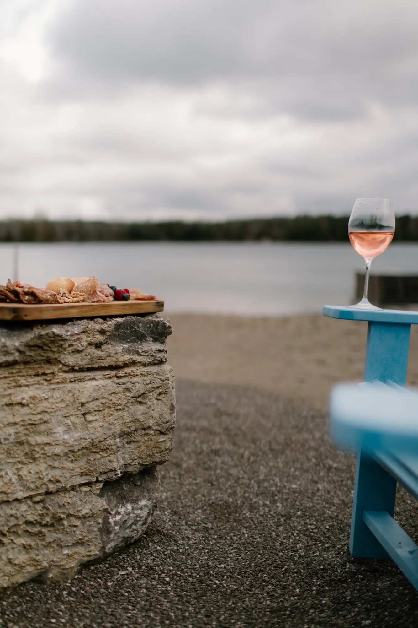 A charcuterie board on a stone ledge and a glass of rosé on the arm of a blue Adirondack chair, with the lake and its far shore behind.