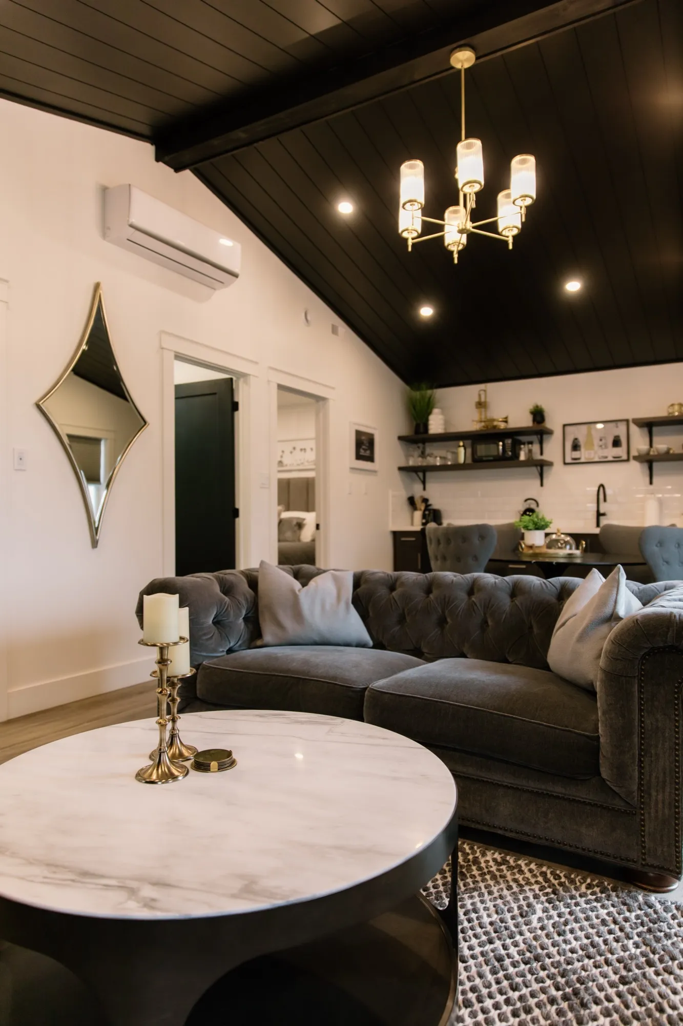 The Vintage Hollywood living area, with a grey tufted sofa, a brass chandelier under a dark plank ceiling, and a marble-topped coffee table.
